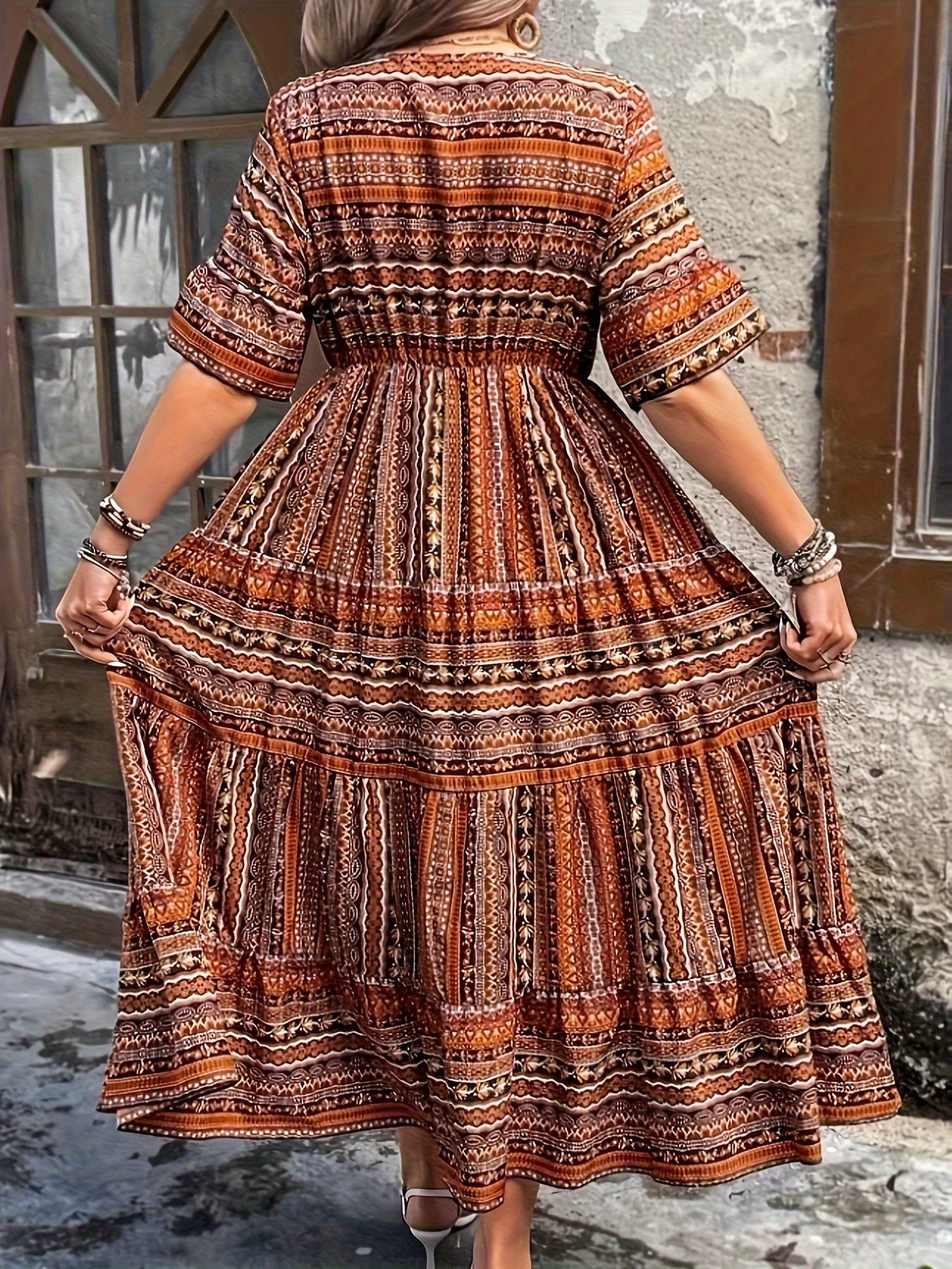 Brown - A woman wearing an olive green bohemian geometric print maxi sundress with a V-neckline, elbow-length sleeves, and a tiered skirt, holding a small woven straw handbag.