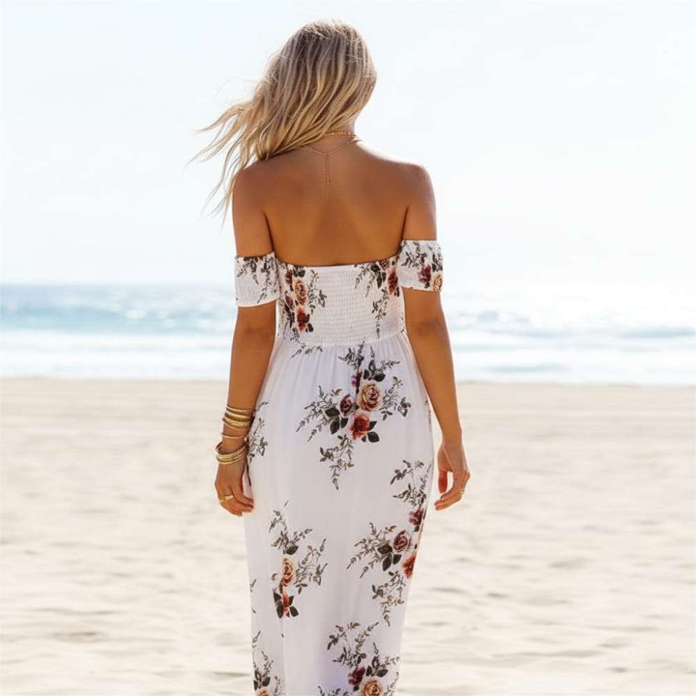 Gray - A woman standing on a sandy beach wearing a white off-the-shoulder chiffon maxi dress with a botanical floral print, featuring a smocked bodice and a high thigh slit.