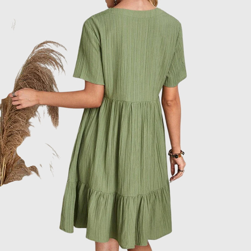 Pink - A woman wearing a dusty rose crinkle gauze short-sleeve V-neck sundress with an empire waist and tiered ruffle hem, styled with a straw shoulder bag.