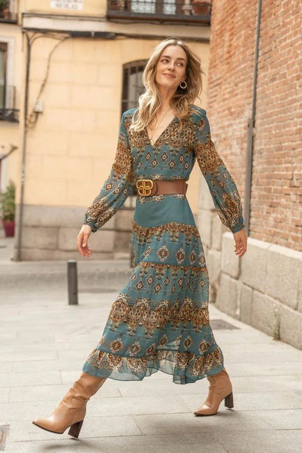 3XL - A woman wearing a teal and brown paisley print chiffon maxi dress with long bishop sleeves, a V-neckline, and a tiered ruffle skirt, styled with a brown leather belt and tan knee-high boots.