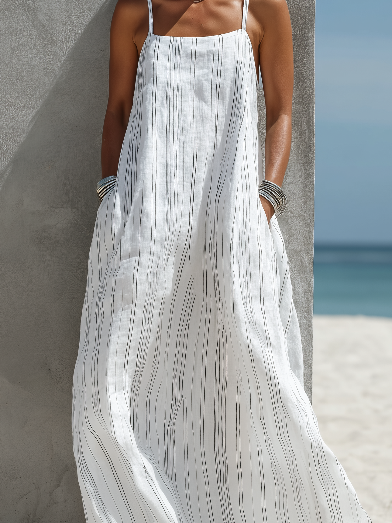 White - Woman standing on a beach wearing a relaxed fit, striped spaghetti-strap maxi dress with her hands in the side pockets.