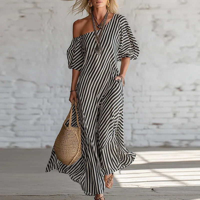 Black - Woman wearing a long, striped, one-shoulder maxi dress with a puff sleeve and a side pocket, holding a woven tote bag.