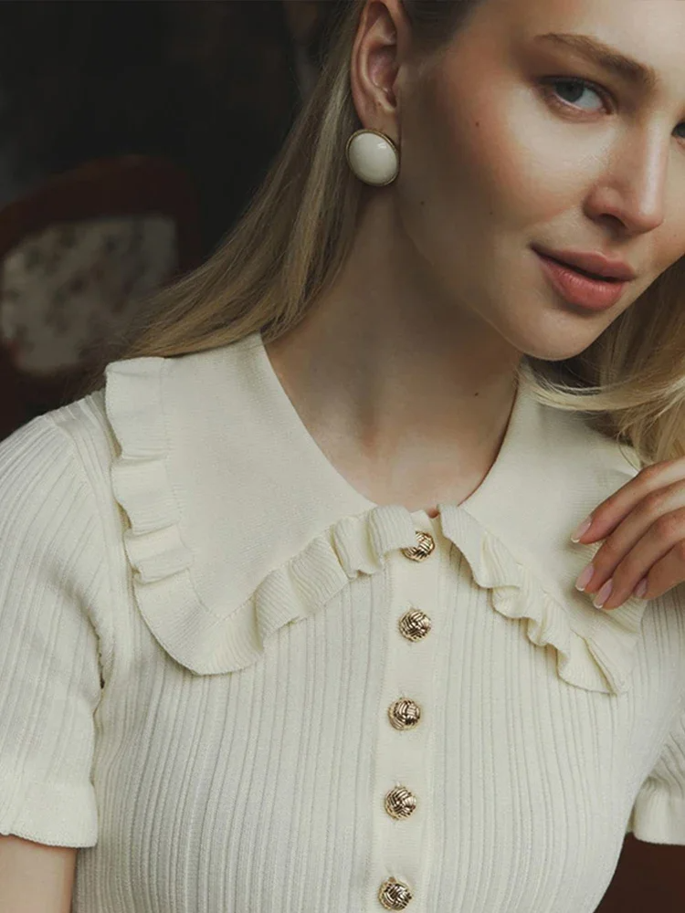 Woman in cream ribbed knit mini dress with ruffle collar and gold buttons sitting at a table.