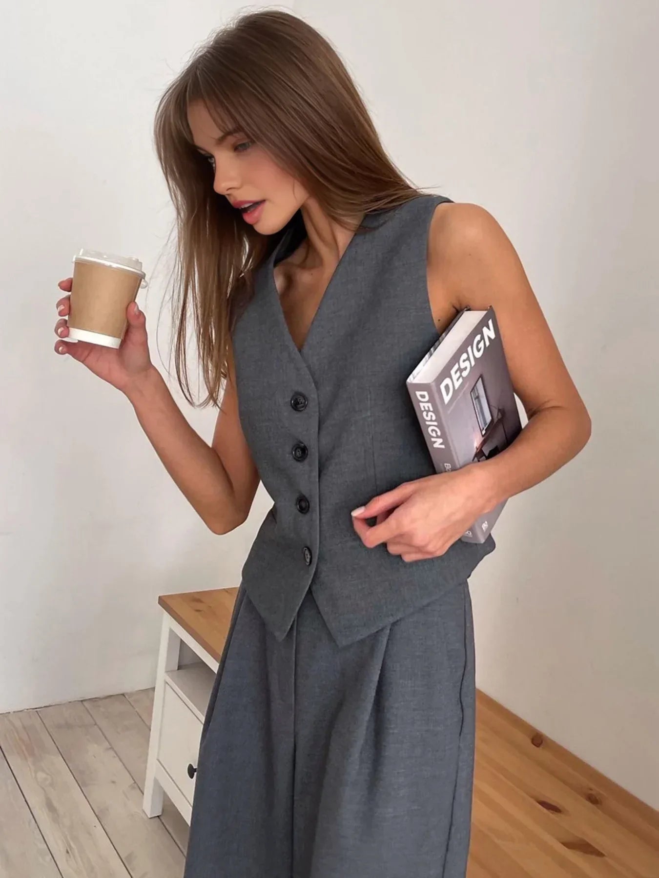 Woman in gray wool blend vest and wide leg pants holding a coffee cup and design book.