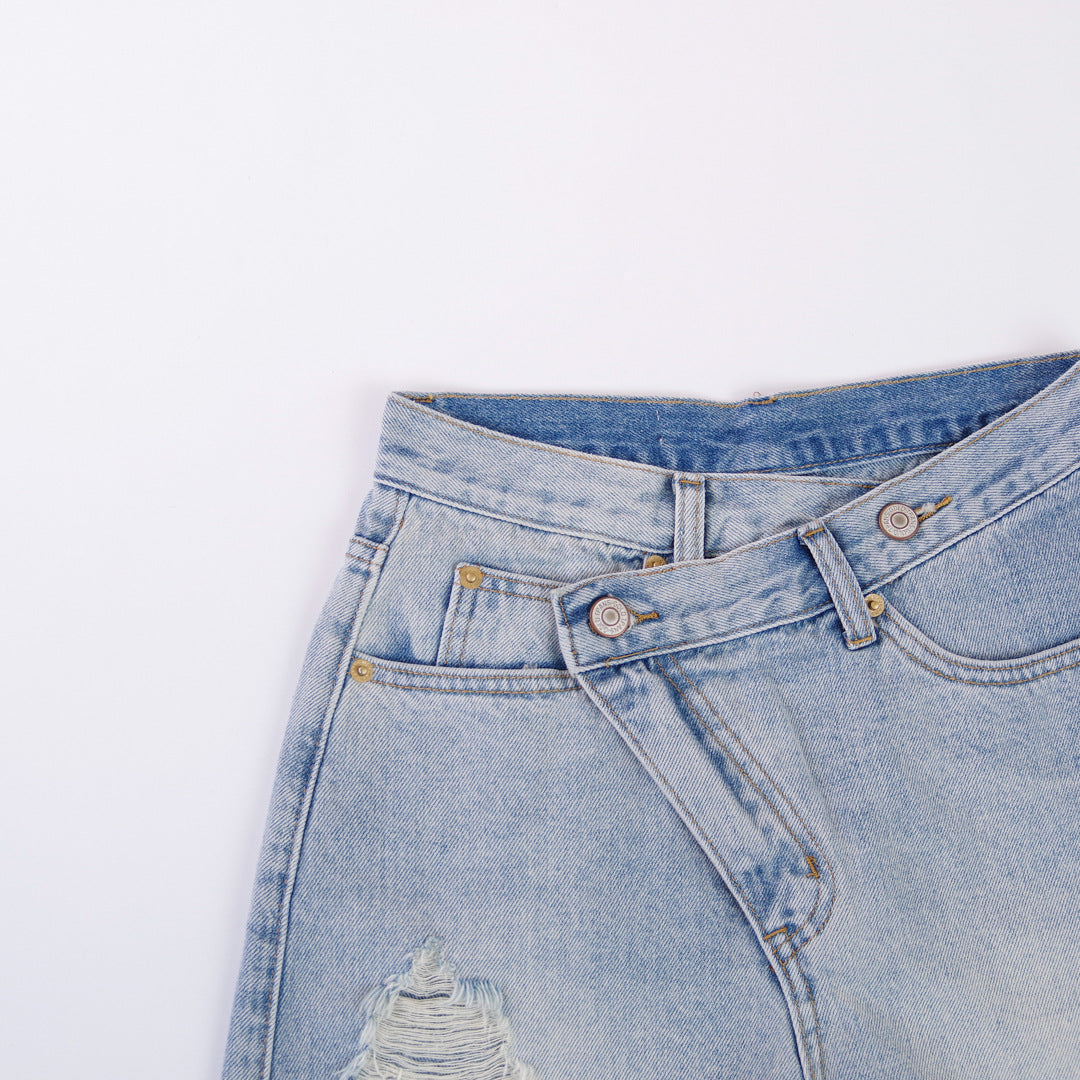 Woman wearing light wash denim shorts with asymmetrical waist and distressed hem.