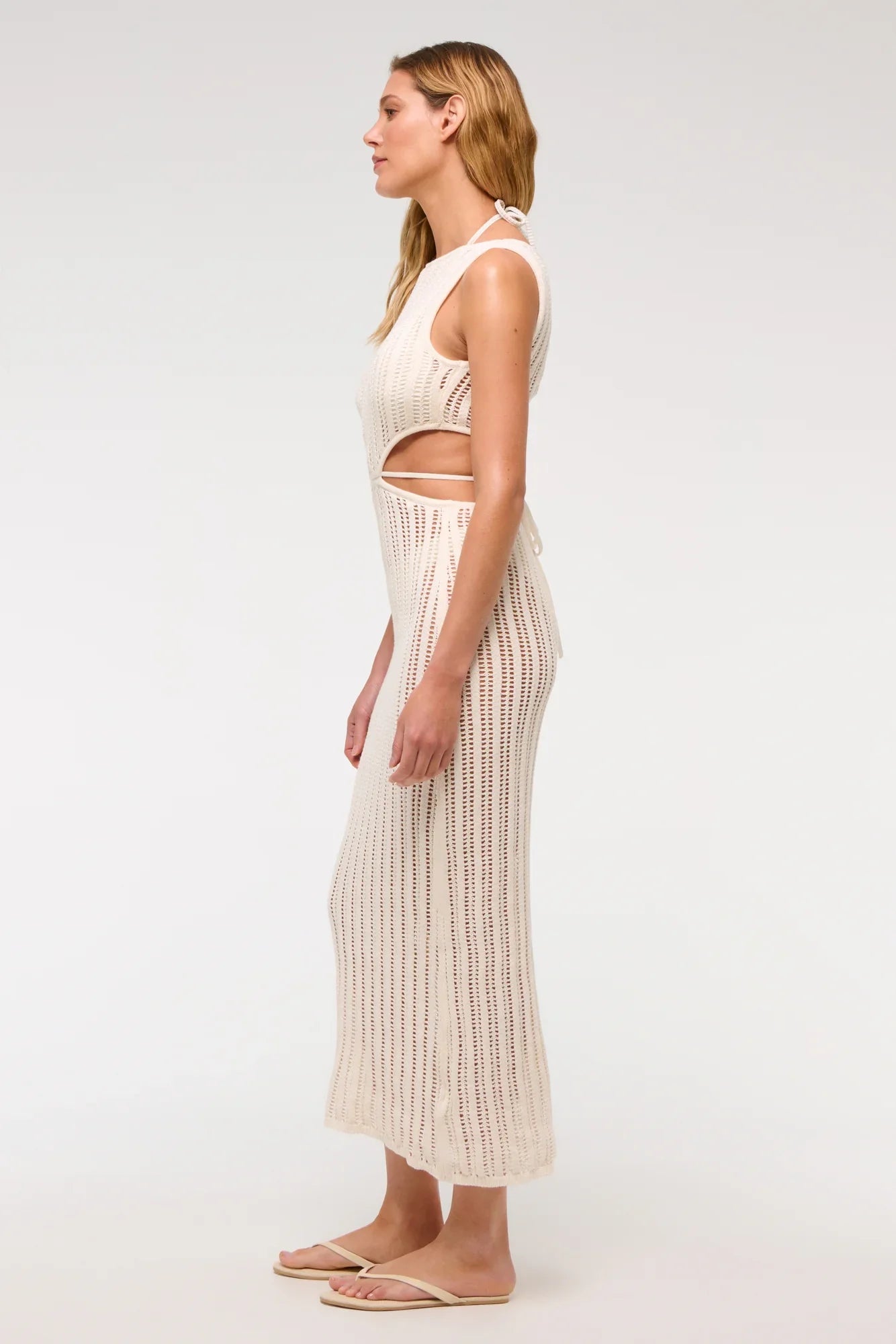 Woman wearing a long, off-white crochet knit dress with waist cutouts.
