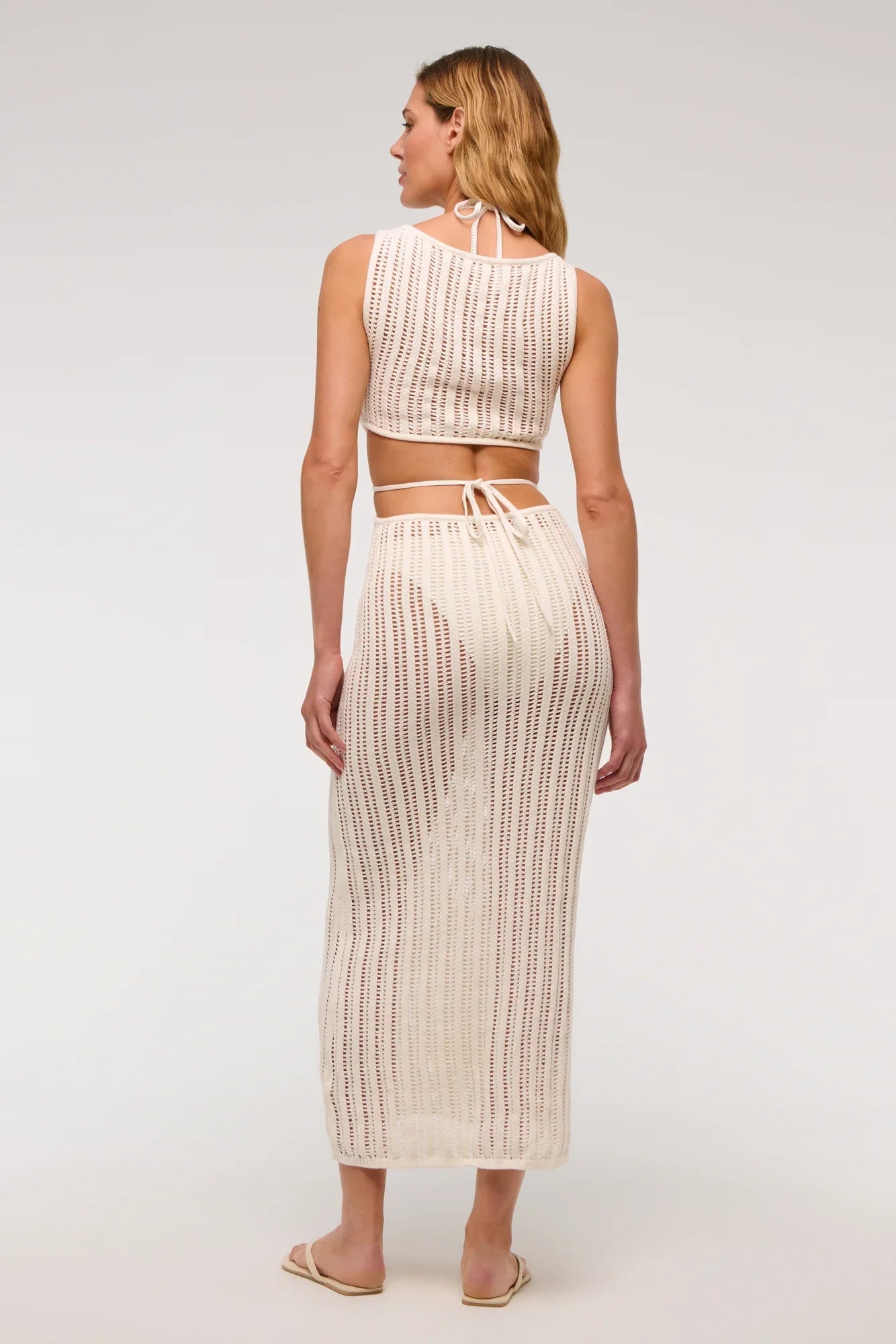 Woman wearing a long, off-white crochet knit dress with waist cutouts.