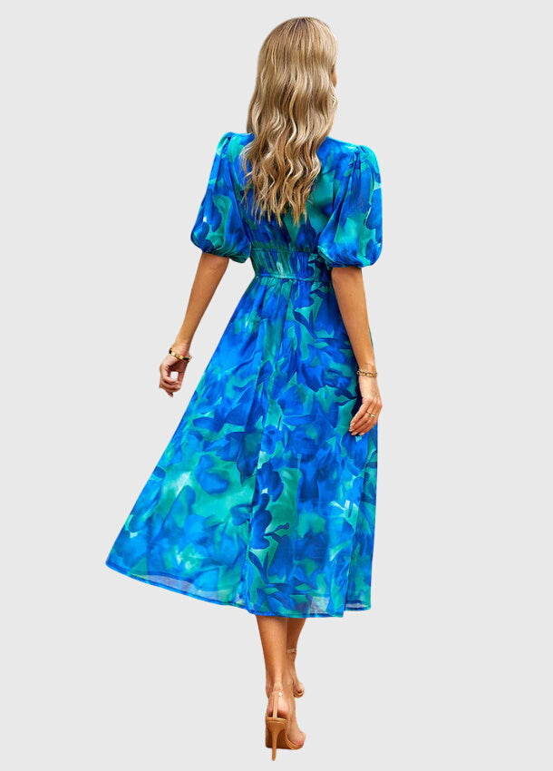 Blue - A woman wearing a vibrant multi-color abstract floral print chiffon midi dress with puff sleeves, a V-neckline, and a smocked waist, paired with white heeled sandals.