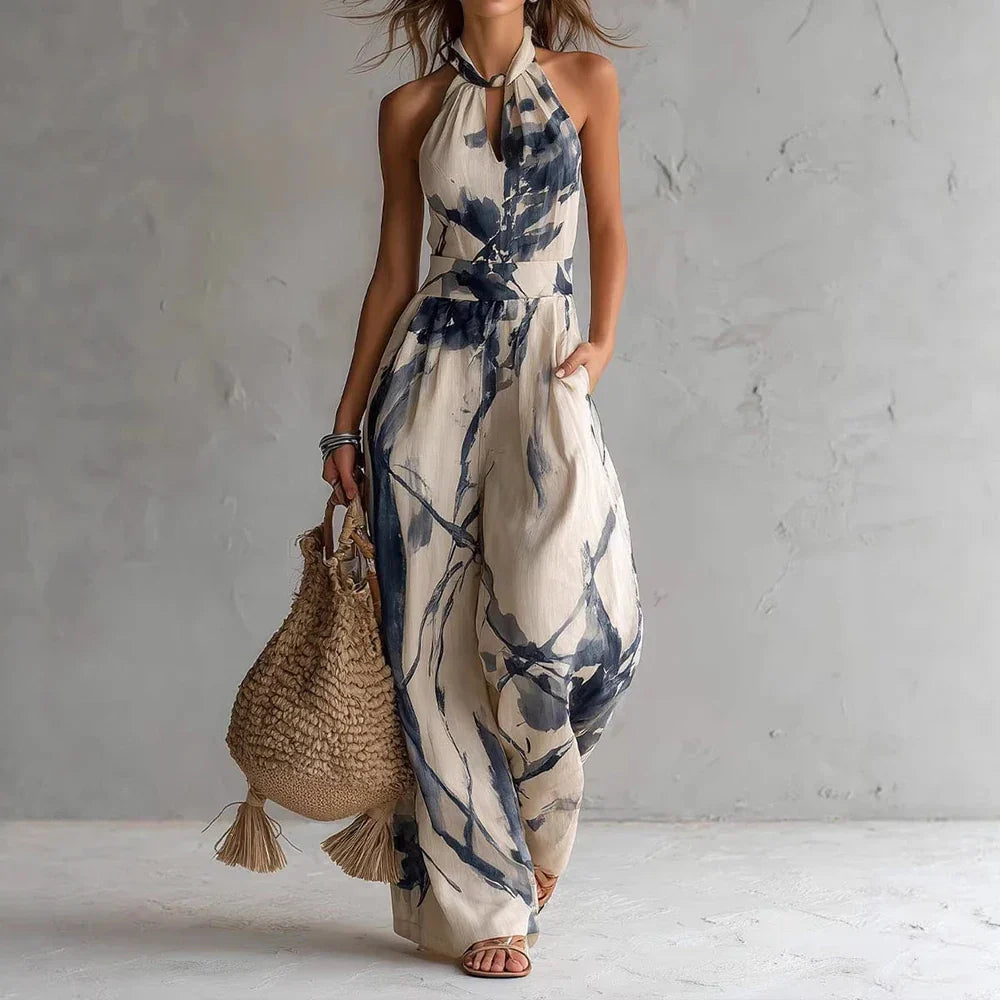Light Blue - A woman wearing a cream-colored, halter-neck wide-leg jumpsuit with a navy blue abstract watercolor print, holding a woven straw bag.