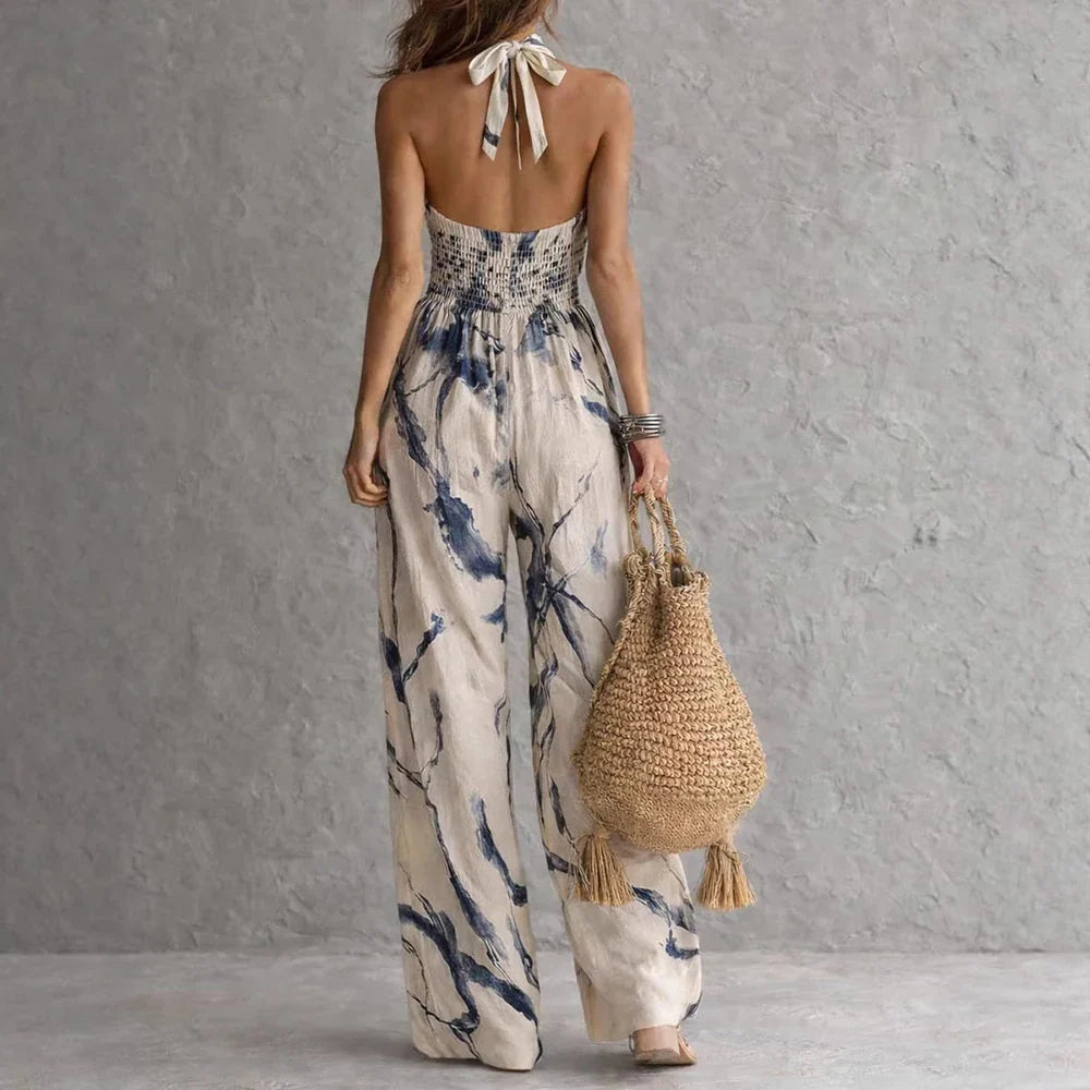 Light Blue - A woman wearing a cream-colored, halter-neck wide-leg jumpsuit with a navy blue abstract watercolor print, holding a woven straw bag.