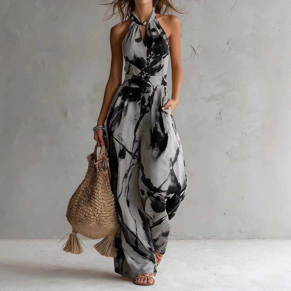 Black - A woman wearing a cream-colored, halter-neck wide-leg jumpsuit with a navy blue abstract watercolor print, holding a woven straw bag.