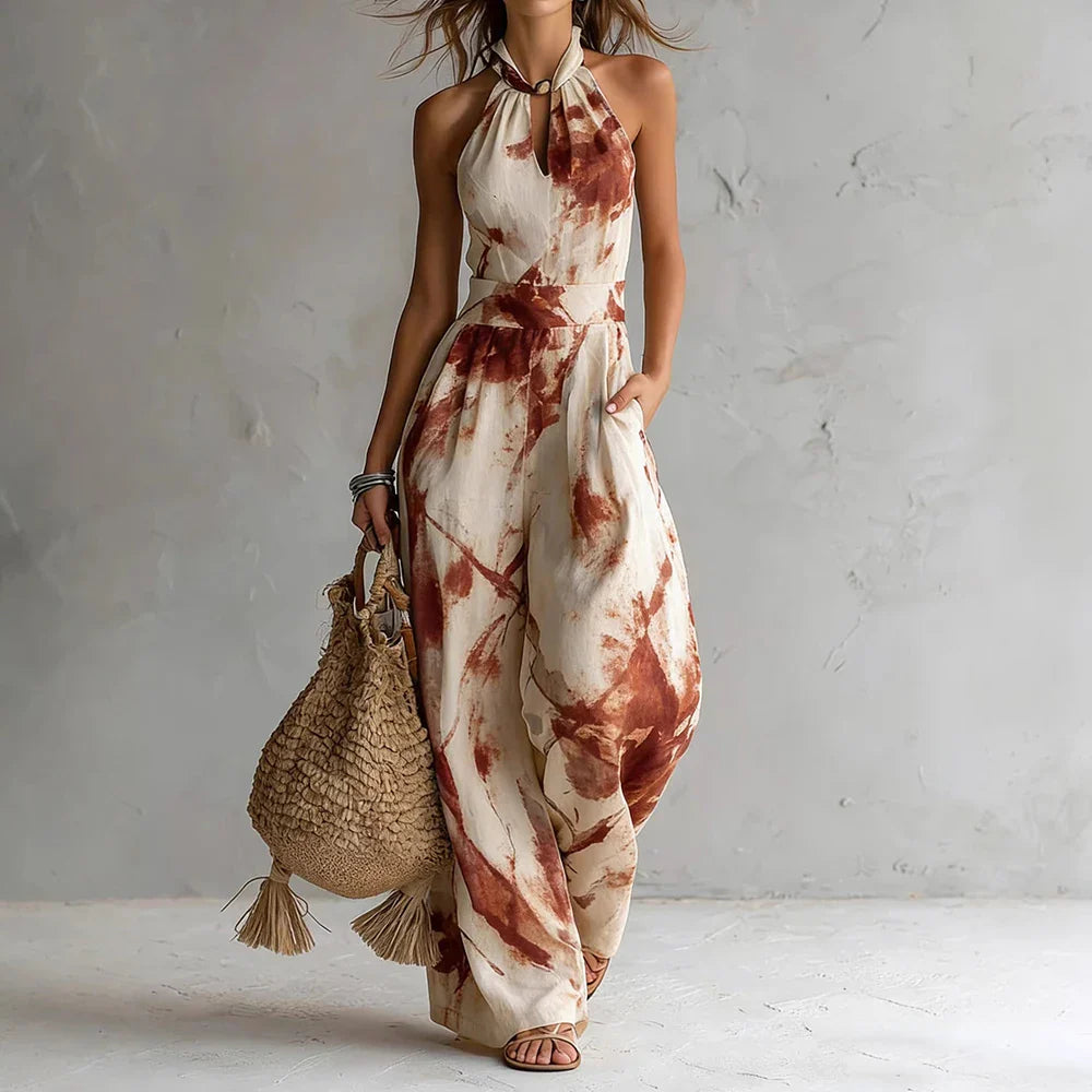 Red - A woman wearing a cream-colored, halter-neck wide-leg jumpsuit with a navy blue abstract watercolor print, holding a woven straw bag.