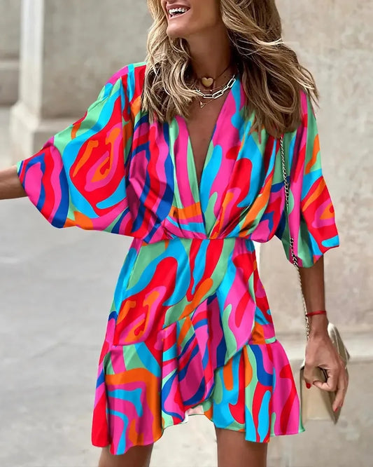 Rose Red - A woman wearing a vibrant, multi-colored abstract print mini dress featuring a deep V-neckline, wide kimono sleeves, and a draped wrap skirt.