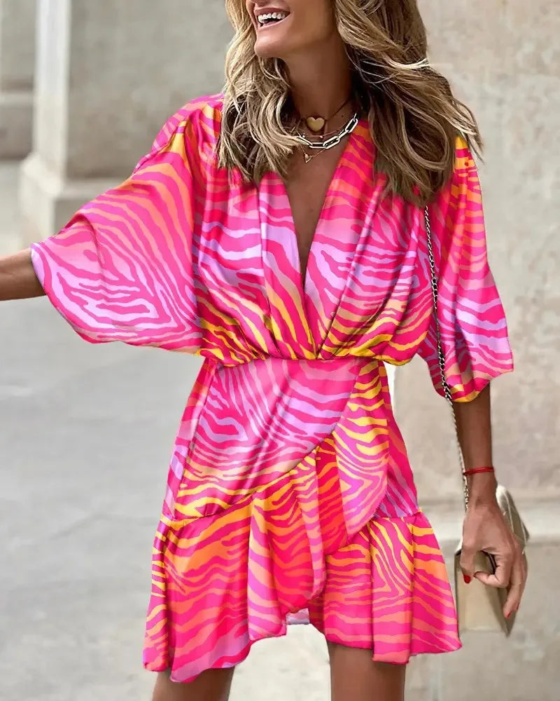Pink - A woman wearing a vibrant, multi-colored abstract print mini dress featuring a deep V-neckline, wide kimono sleeves, and a draped wrap skirt.