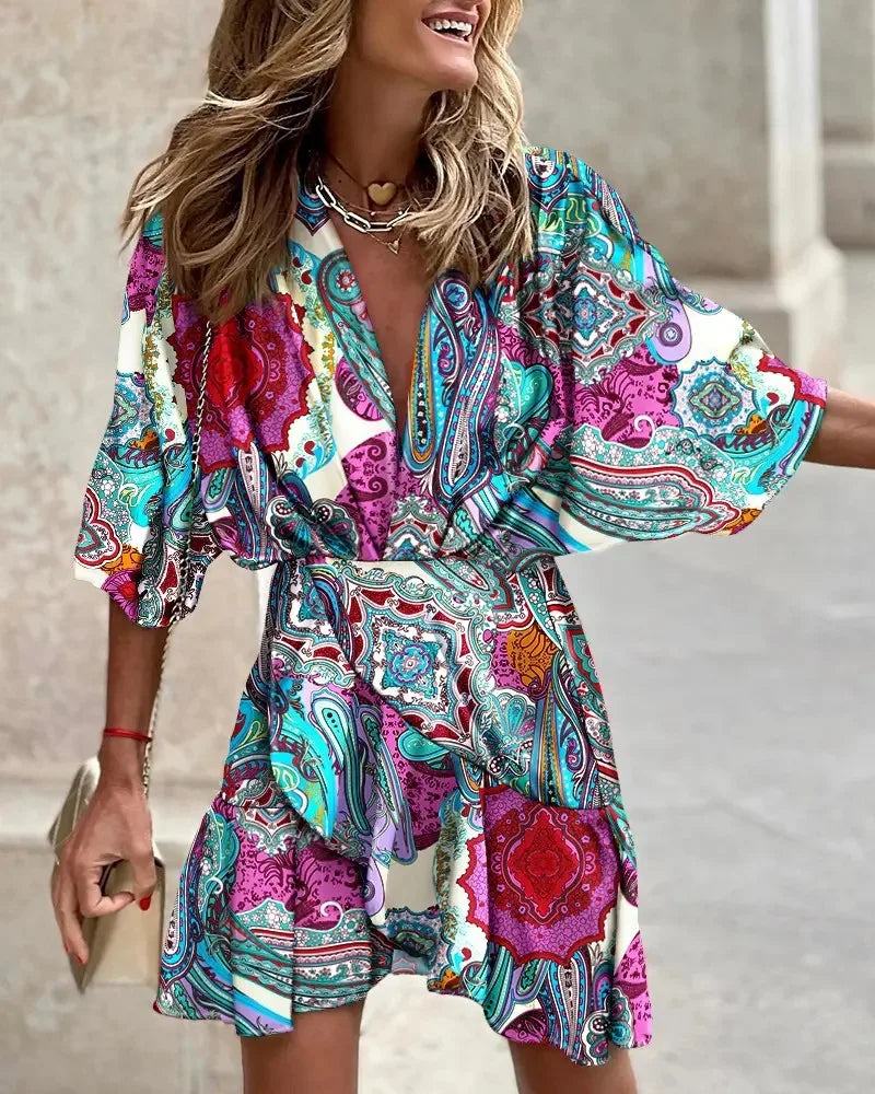 Blue - A woman wearing a vibrant, multi-colored abstract print mini dress featuring a deep V-neckline, wide kimono sleeves, and a draped wrap skirt.