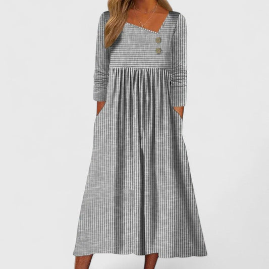 Gray - A woman wearing a gray and white vertically striped midi sundress with three-quarter sleeves, an asymmetric neckline featuring two decorative buttons, an empire waist, and side pockets.