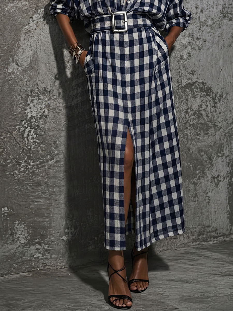 Blue And White - Model wearing a long plaid button-down maxi shirtdress with a wide matching belt, front slit, and rolled sleeves, leaning against a textured wall.
