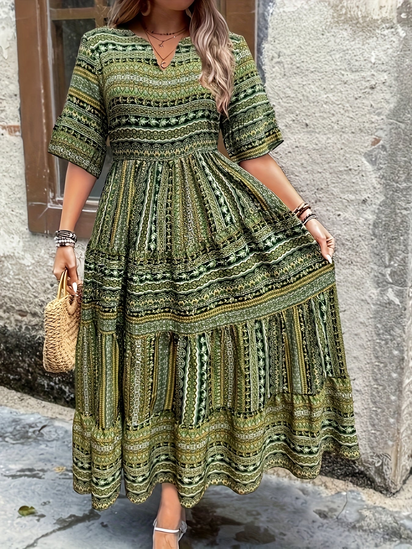A woman wearing an olive green bohemian geometric print maxi sundress with a V-neckline, elbow-length sleeves, and a tiered skirt, holding a small woven straw handbag.