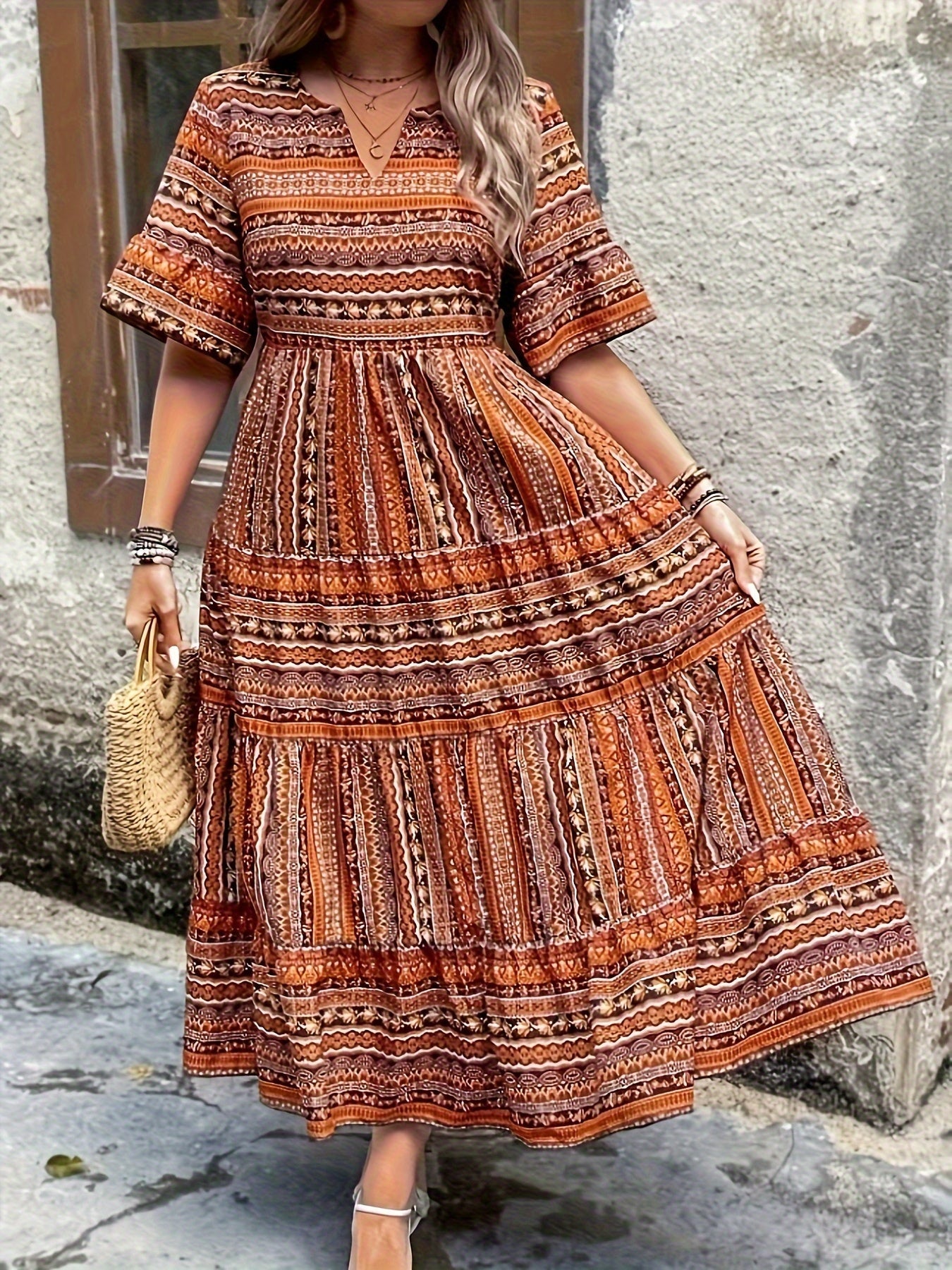 Brown - A woman wearing an olive green bohemian geometric print maxi sundress with a V-neckline, elbow-length sleeves, and a tiered skirt, holding a small woven straw handbag.