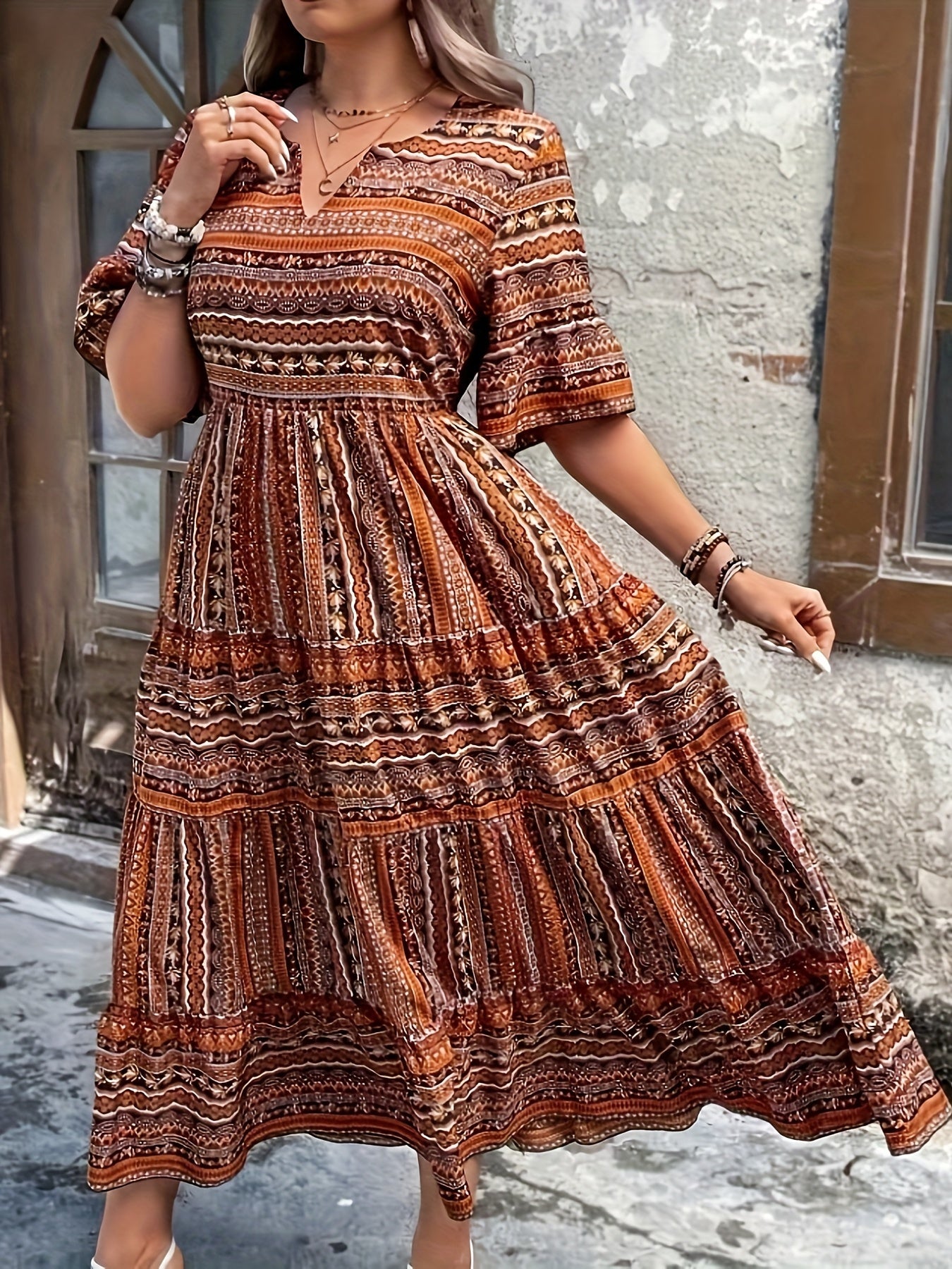 Brown - A woman wearing an olive green bohemian geometric print maxi sundress with a V-neckline, elbow-length sleeves, and a tiered skirt, holding a small woven straw handbag.