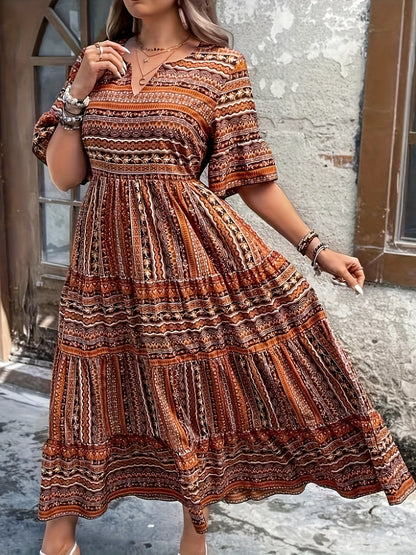 Brown - A woman wearing an olive green bohemian geometric print maxi sundress with a V-neckline, elbow-length sleeves, and a tiered skirt, holding a small woven straw handbag.