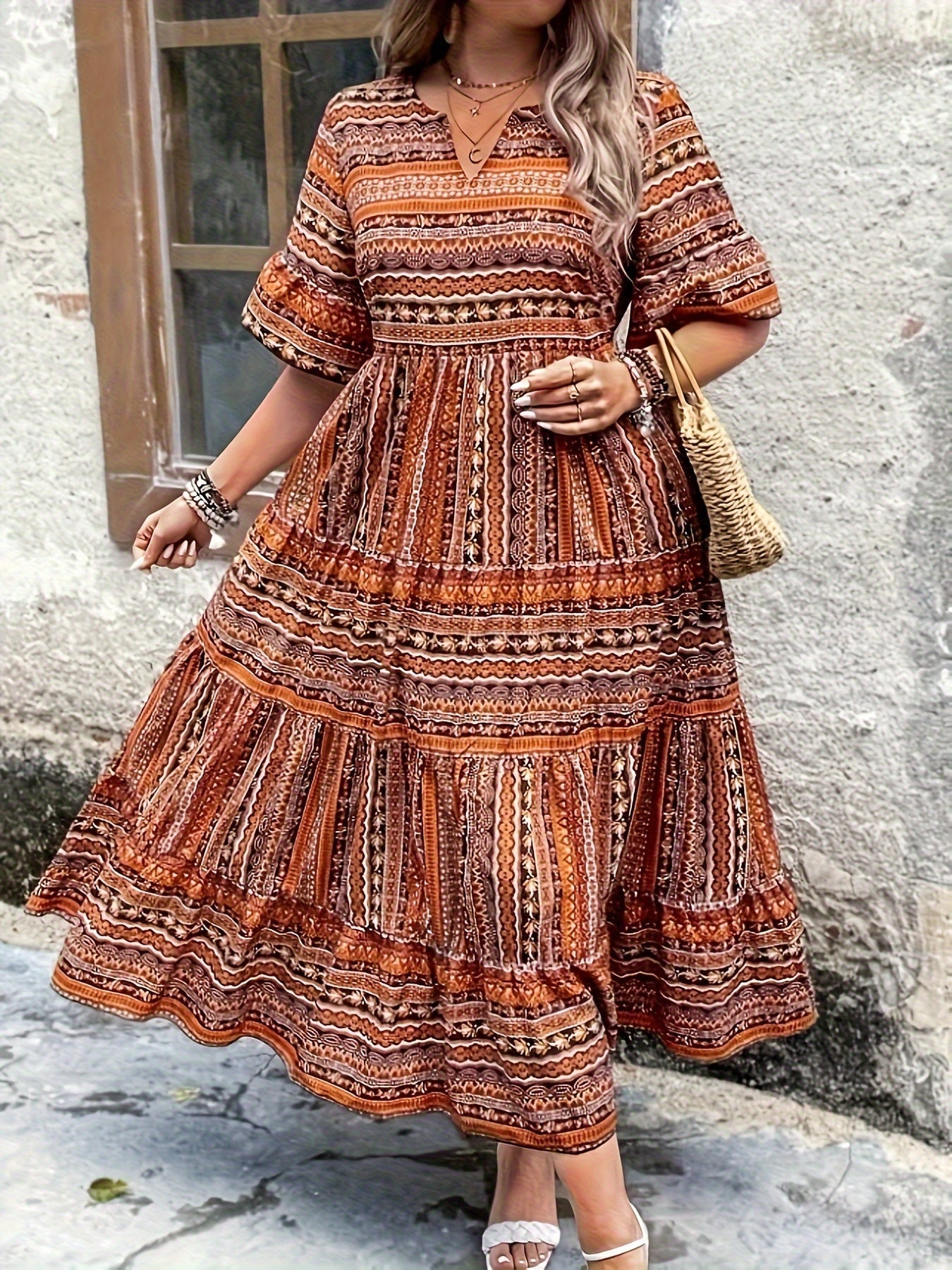 Brown - A woman wearing an olive green bohemian geometric print maxi sundress with a V-neckline, elbow-length sleeves, and a tiered skirt, holding a small woven straw handbag.