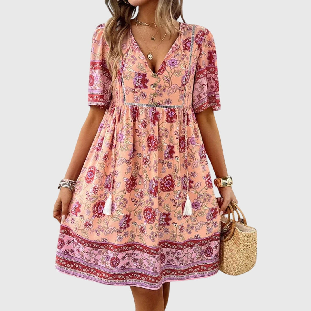 Pink - A woman wearing a peach-colored bohemian mini sundress featuring a pink and burgundy paisley floral print, short sleeves, a V-neckline with tassel ties, and a border print hem.