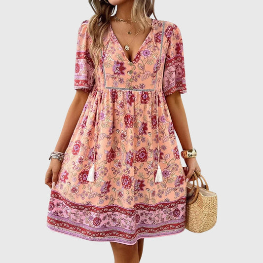 Pink - A woman wearing a peach-colored bohemian mini sundress featuring a pink and burgundy paisley floral print, short sleeves, a V-neckline with tassel ties, and a border print hem.