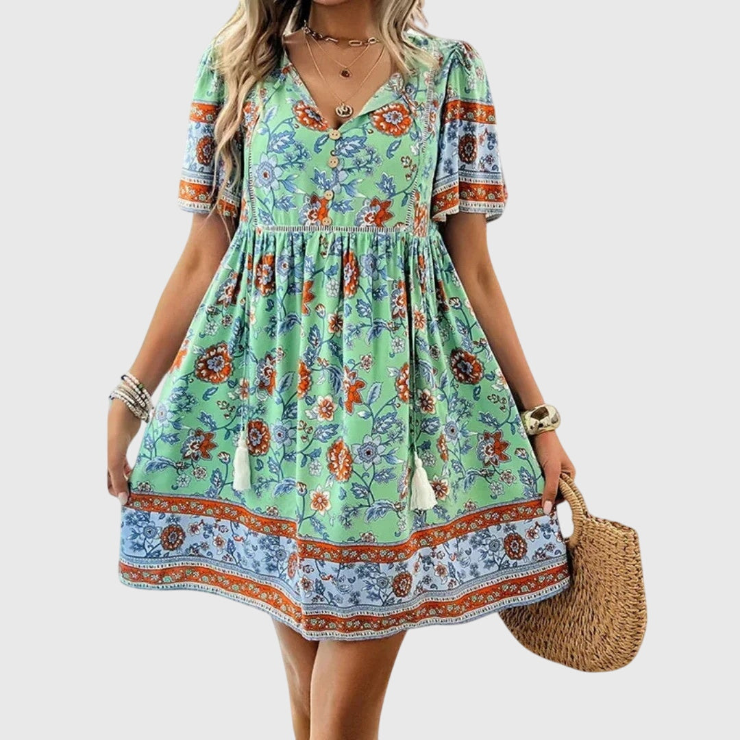 Green - A woman wearing a peach-colored bohemian mini sundress featuring a pink and burgundy paisley floral print, short sleeves, a V-neckline with tassel ties, and a border print hem.