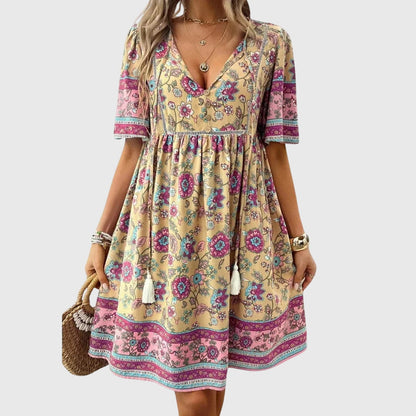 Beige - A woman wearing a peach-colored bohemian mini sundress featuring a pink and burgundy paisley floral print, short sleeves, a V-neckline with tassel ties, and a border print hem.