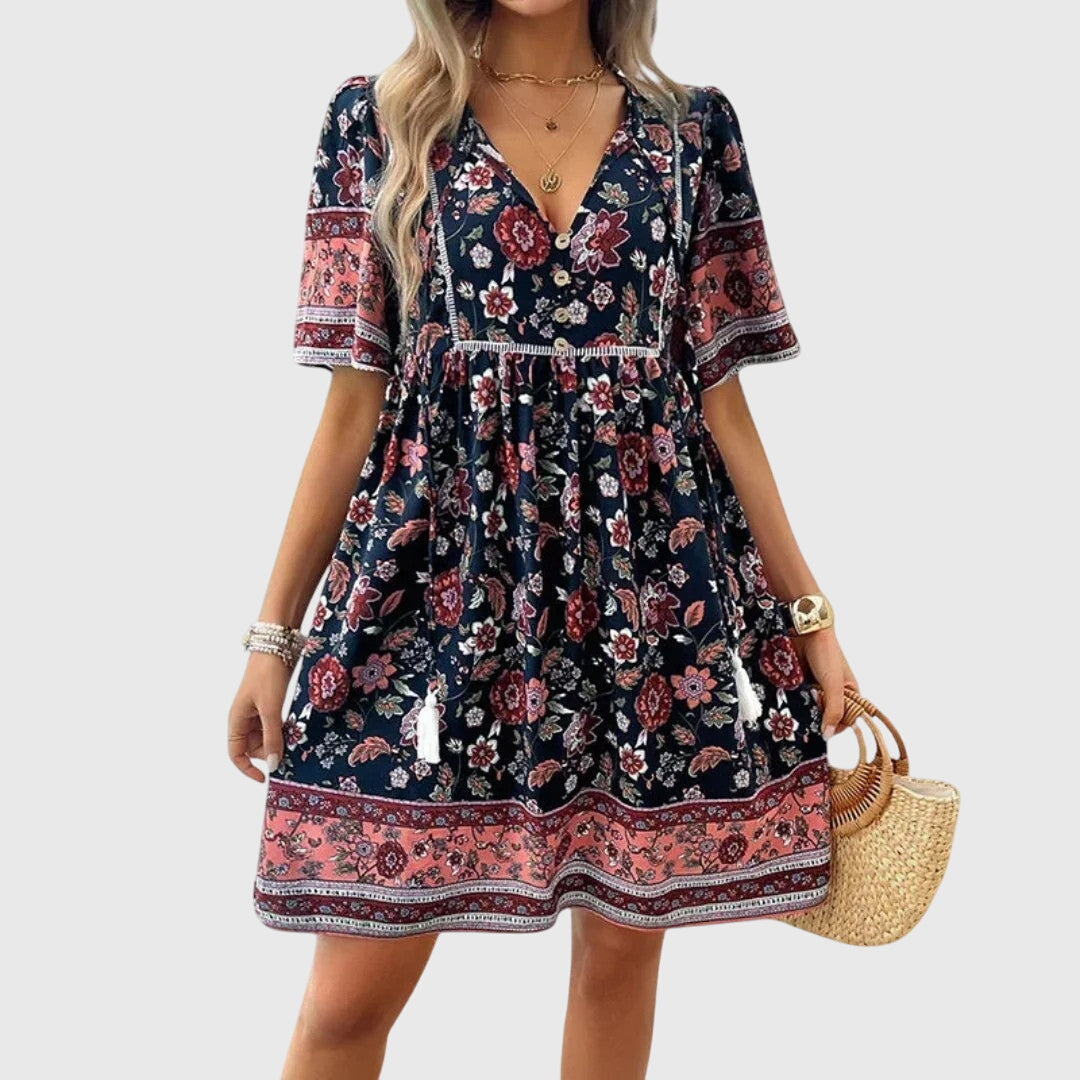 Black - A woman wearing a peach-colored bohemian mini sundress featuring a pink and burgundy paisley floral print, short sleeves, a V-neckline with tassel ties, and a border print hem.