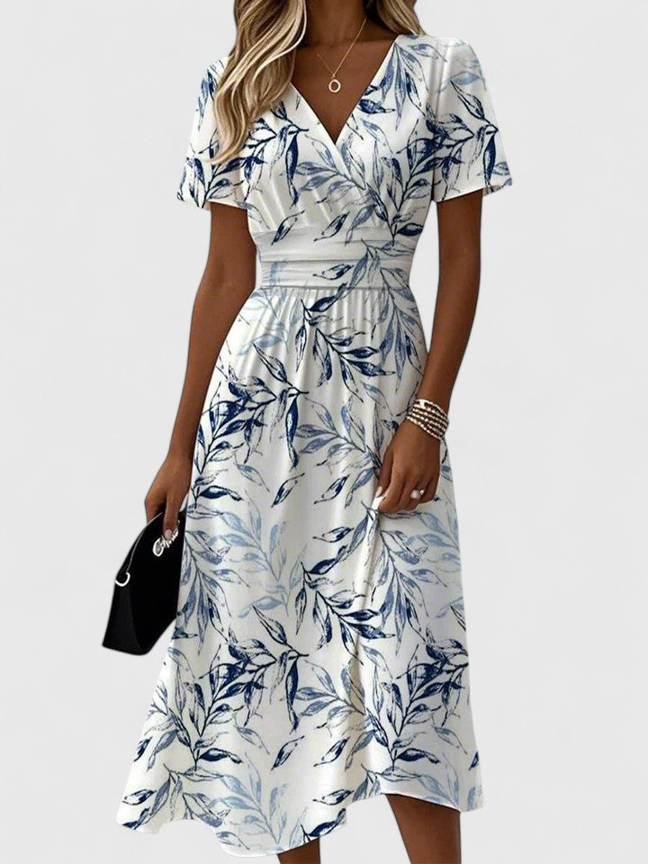 White - A woman wearing a white midi dress featuring a blue botanical leaf print, a surplice V-neckline, flutter sleeves, and a defined banded waist.
