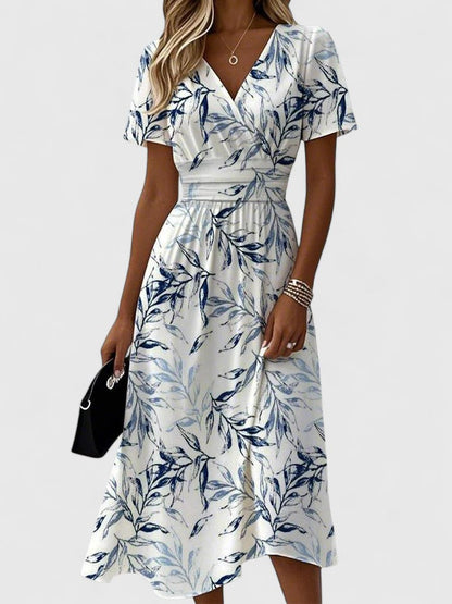 White - A woman wearing a white midi dress featuring a blue botanical leaf print, a surplice V-neckline, flutter sleeves, and a defined banded waist.