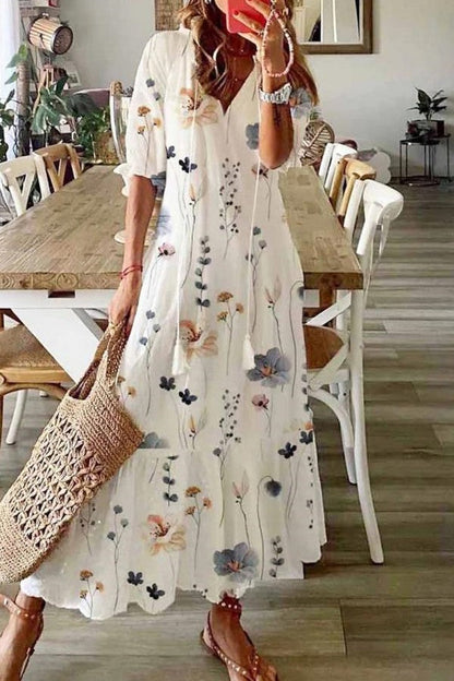 Floral White - A woman wearing a white short-sleeve maxi sundress with a colorful botanical wildflower print, featuring a V-neckline with tassel ties and a tiered hem, paired with a woven straw tote bag.