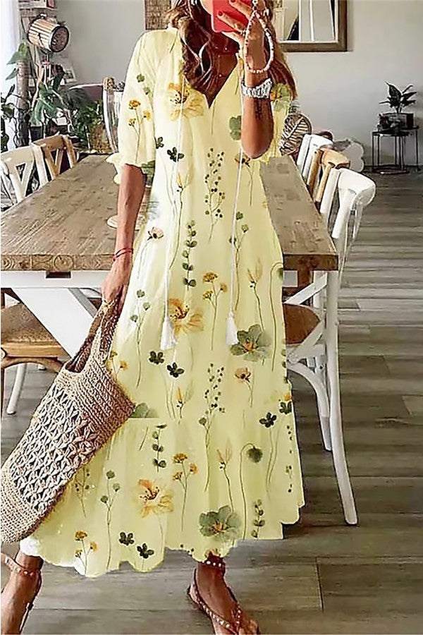 Yellow - A woman wearing a white short-sleeve maxi sundress with a colorful botanical wildflower print, featuring a V-neckline with tassel ties and a tiered hem, paired with a woven straw tote bag.