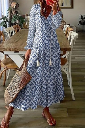 Blue - A woman wearing a white short-sleeve maxi sundress with a colorful botanical wildflower print, featuring a V-neckline with tassel ties and a tiered hem, paired with a woven straw tote bag.
