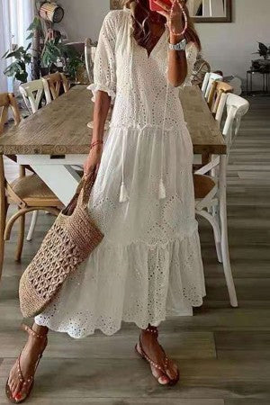 White - A woman wearing a white short-sleeve maxi sundress with a colorful botanical wildflower print, featuring a V-neckline with tassel ties and a tiered hem, paired with a woven straw tote bag.