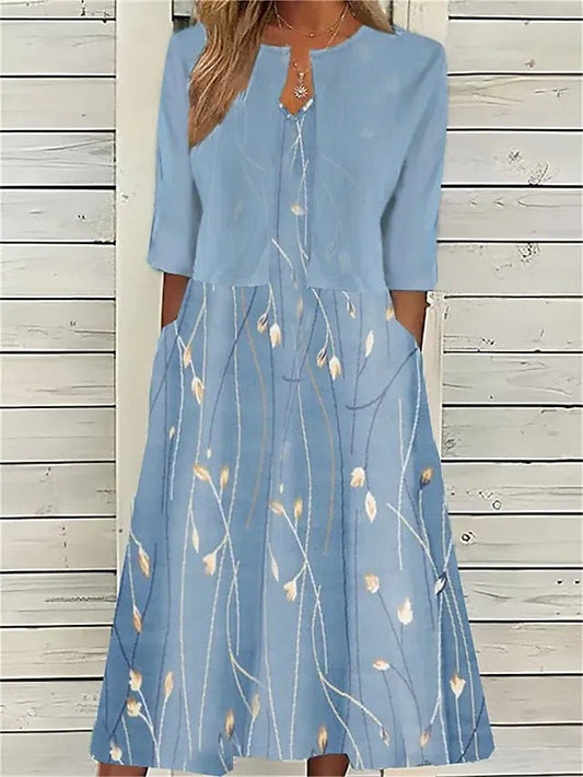 A woman wearing a dusty blue mock-layered midi dress featuring a solid 3/4-sleeve bodice, a split neckline, and a flowy skirt with a white botanical branch print.