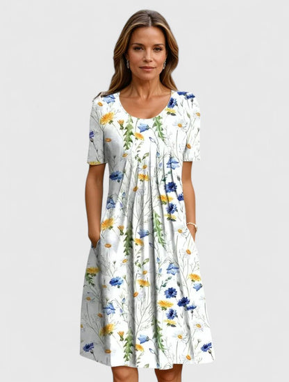 Yellow Flowers - A woman wearing a white short-sleeve A-line sundress featuring a vibrant yellow and blue wildflower botanical print, complete with a scoop neckline and side pockets.