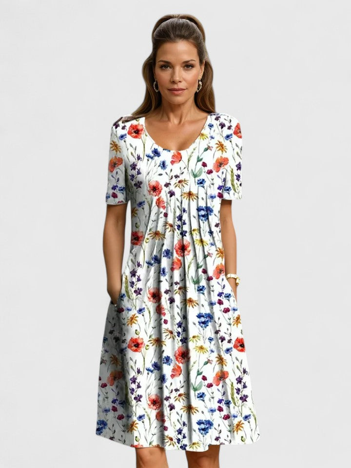 Red Flowers - A woman wearing a white short-sleeve A-line sundress featuring a vibrant yellow and blue wildflower botanical print, complete with a scoop neckline and side pockets.
