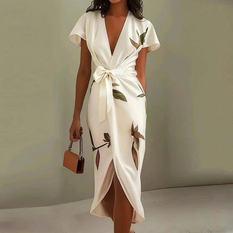 White - A woman wearing a cream-colored satin midi dress featuring a green botanical leaf print, a deep V-neckline, flutter sleeves, a tie-front waist bow, and a wrap-style tulip hem.