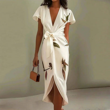 White - A woman wearing a cream-colored satin midi dress featuring a green botanical leaf print, a deep V-neckline, flutter sleeves, a tie-front waist bow, and a wrap-style tulip hem.