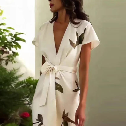White - A woman wearing a cream-colored satin midi dress featuring a green botanical leaf print, a deep V-neckline, flutter sleeves, a tie-front waist bow, and a wrap-style tulip hem.