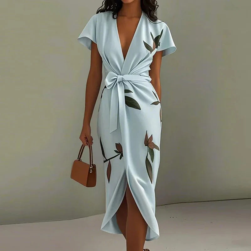 Sky Blue - A woman wearing a cream-colored satin midi dress featuring a green botanical leaf print, a deep V-neckline, flutter sleeves, a tie-front waist bow, and a wrap-style tulip hem.