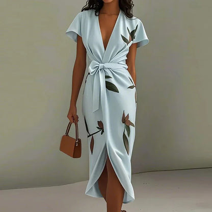 Sky Blue - A woman wearing a cream-colored satin midi dress featuring a green botanical leaf print, a deep V-neckline, flutter sleeves, a tie-front waist bow, and a wrap-style tulip hem.