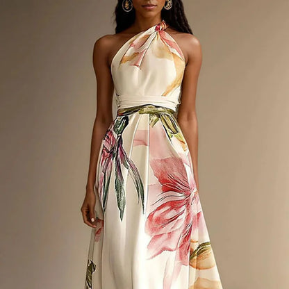 Cream - A woman wearing a floor-length satin halter maxi dress featuring a large-scale watercolor botanical print in pink, orange, and green on an ivory background, showcasing a pleated waist and a flowing A-line silhouette.