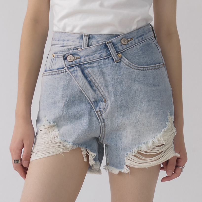 Woman wearing light wash denim shorts with asymmetrical waist and distressed hem.