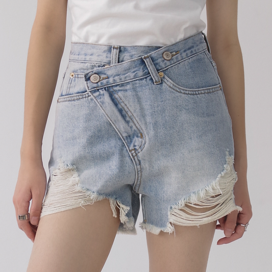 Woman wearing light wash denim shorts with asymmetrical waist and distressed hem.
