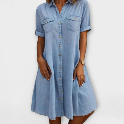 Blue - A woman wearing a light blue chambray short-sleeve button-down sundress with chest pockets and a relaxed fit.