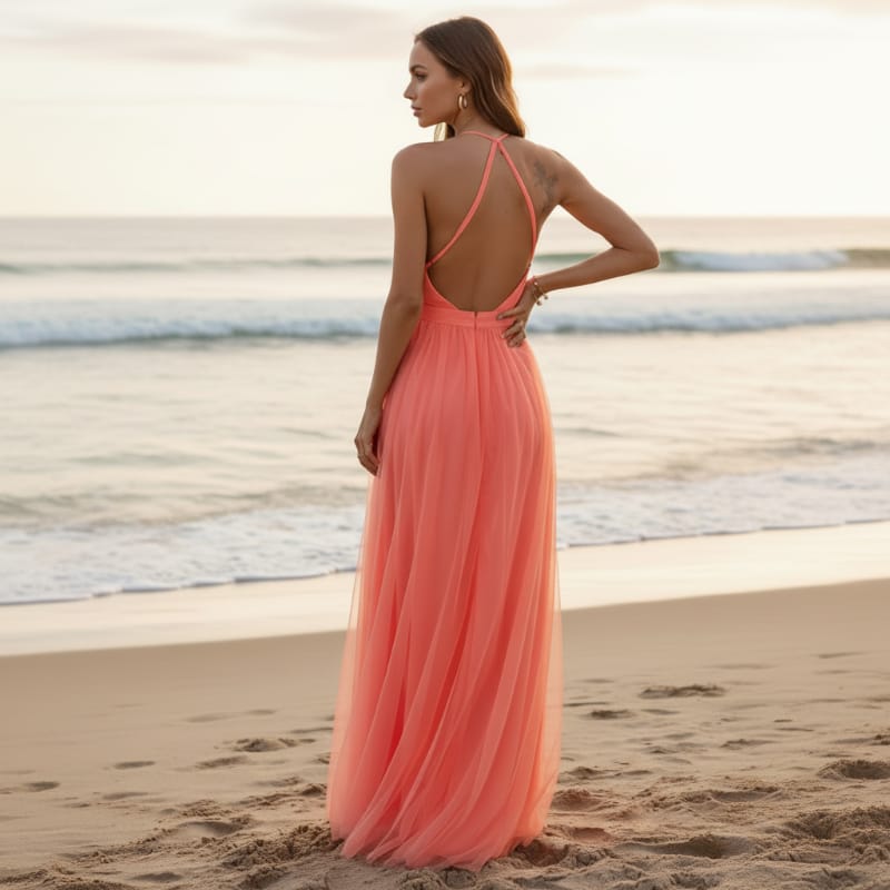 Orange - A woman stands on a sandy beach at sunset wearing a floor-length coral chiffon halter dress with a plunging V-neckline and a flowing, layered maxi skirt.